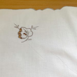 May include: A white rectangular textile featuring a scalloped edge with light gray embroidery. The fabric displays a delicate floral motif created with cutwork and gray thread stitching, positioned near the corner of the piece.