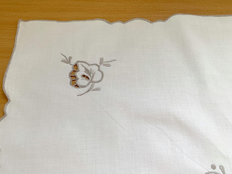 May include: A white rectangular textile featuring a scalloped edge with light gray embroidery. The fabric displays a delicate floral motif created with cutwork and gray thread stitching, positioned near the corner of the piece.