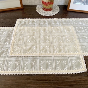 Può includere: Due tovagliette rettangolari con un motivo a griglia color beige chiaro e bianco sporco. Ogni tovaglietta è decorata con motivi ricamati bianchi a forma di trifoglio all'interno di ogni quadrato e rifinita con un bordo smerlato in tinta.