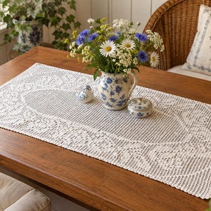 May include: A rectangular white crochet table runner featuring an intricate openwork lace pattern with floral motifs. The runner is placed on a wooden surface and holds a white ceramic pitcher with blue floral patterns, a small bird figurine, and a lidded jar.
