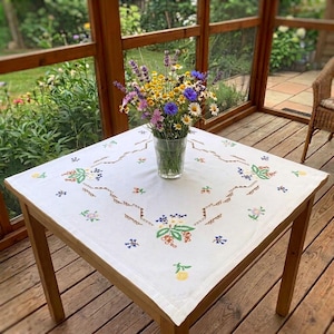 Peut inclure: Une nappe carrée blanche ornée de motifs floraux brodés en vert, bleu, jaune et rouge, surmontée d'un vase en verre transparent contenant un bouquet de fleurs sauvages fraîches, notamment de la lavande violette, des marguerites et des bleuets.