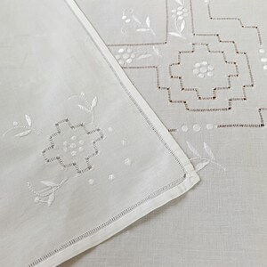May include: A white linen textile featuring intricate white embroidery with geometric patterns and floral motifs. The fabric displays delicate cutwork details along the edges and a hemstitched border, showcasing a clean and crisp appearance.