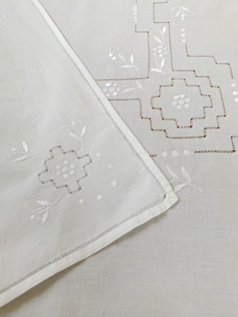 May include: A white linen textile featuring intricate white embroidery with geometric patterns and floral motifs. The fabric displays delicate cutwork details along the edges and a hemstitched border, showcasing a clean and crisp appearance.