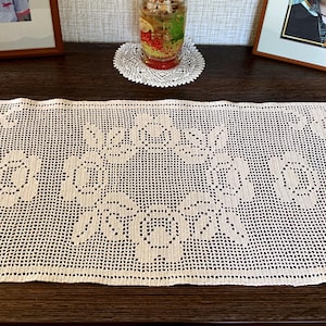 May include: A rectangular off-white crochet table runner featuring a repeating openwork floral pattern. The design consists of interconnected roses and leaves created with a grid-like stitch technique, forming a symmetrical arrangement across the fabric.