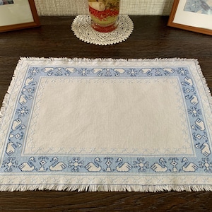 May include: A rectangular placemat with a light beige center and a light blue border featuring a repeating cross-stitch pattern of white swans and geometric floral motifs. The edges of the fabric are finished with a short, frayed fringe.