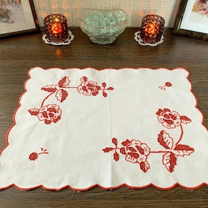 Pode incluir: Um lugar americano retangular de tecido branco com bordado de flores em ponto cruz vermelho. As bordas são recortadas e finalizadas com linha vermelha. O design inclui dois grupos florais principais com caules e folhas nos cantos opostos.