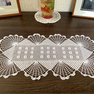 May include: A white, handmade, crocheted table runner. The rectangular runner features a detailed openwork design with a central pattern of small squares and fan-shaped edges. The runner is placed on a dark wooden surface.