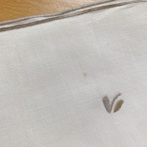 May include: A white linen napkin featuring a light beige embroidered V-shaped monogram in the corner and a matching beige stitched border along the top edge. The fabric has a visible weave texture and a small, faint light brown spot near the center.