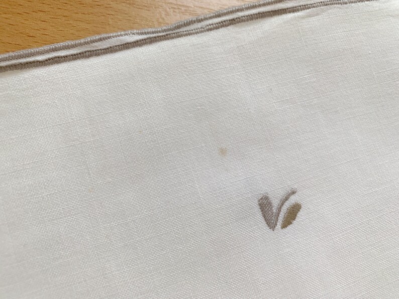 May include: A white linen napkin featuring a light beige embroidered V-shaped monogram in the corner and a matching beige stitched border along the top edge. The fabric has a visible weave texture and a small, faint light brown spot near the center.