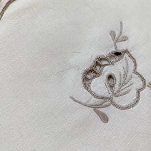 May include: An off-white fabric placemat featuring a scalloped edge with taupe embroidery. The center of the placemat displays a floral motif created with taupe thread, incorporating cutwork details and delicate stitching along the petals and leaves.