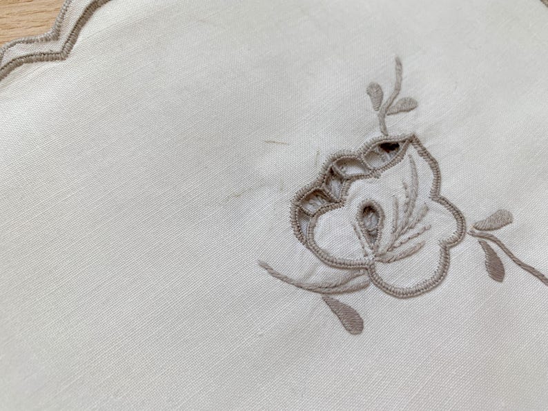 May include: An off-white fabric placemat featuring a scalloped edge with taupe embroidery. The center of the placemat displays a floral motif created with taupe thread, incorporating cutwork details and delicate stitching along the petals and leaves.
