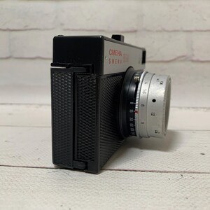 May include: A vintage Smena 8M camera with a silver lens and black body. The camera has the text "CMEHA 8M SMENA" printed in red. The camera is on a light wood surface.