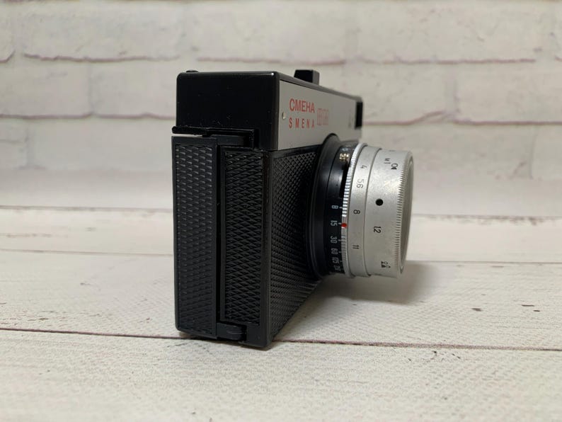May include: A vintage Smena 8M camera with a silver lens and black body. The camera has the text "CMEHA 8M SMENA" printed in red. The camera is on a light wood surface.