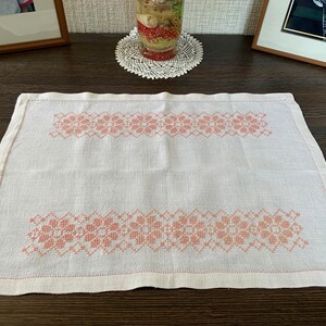May include: A rectangular white linen placemat featuring two horizontal bands of peach-colored cross-stitch embroidery. The embroidery consists of repeating floral patterns and geometric borders. The fabric has a visible weave and a hemmed edge.