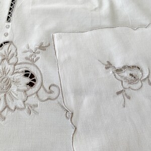 May include: A white textile piece featuring intricate beige floral embroidery and cutwork details. The fabric displays a large, detailed flower motif on the left and a smaller, matching floral accent on the right, with decorative openwork borders.