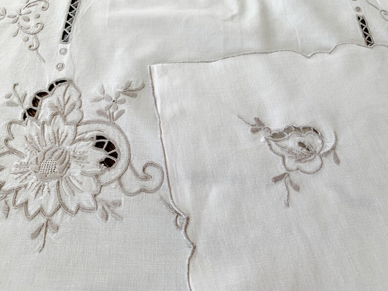 May include: A white textile piece featuring intricate beige floral embroidery and cutwork details. The fabric displays a large, detailed flower motif on the left and a smaller, matching floral accent on the right, with decorative openwork borders.