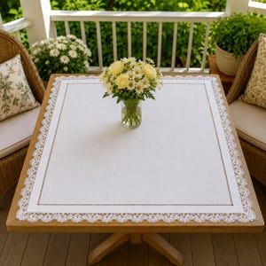 Puede incluir: Un mantel rectangular blanco que presenta un borde de encaje blanco delicado e intrincado con un acabado festoneado. El centro de la tela es blanco liso, enmarcado por una línea de costura fina y sutil. Un florero de cristal contiene un ramo de flores amarillas y blancas.