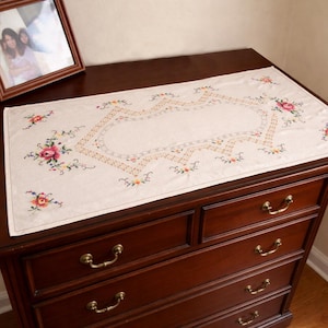 Pode incluir: Um caminho de mesa retangular em tom branco sujo com bordados complexos em ponto cruz. O design inclui um padrão de borda geométrica central em tons de bege e castanho, acentuado por motivos florais coloridos em rosa, amarelo e verde.