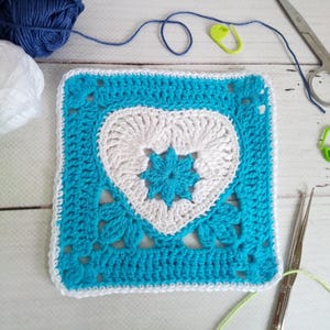 May include: A square, crocheted piece with a white heart design on a turquoise background. The crochet piece is surrounded by white yarn, with balls of pink, white, and blue yarn in the background. Scissors, a crochet hook, and stitch markers are also visible.