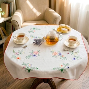 May include: A white square tablecloth with a red scalloped edge, featuring embroidered floral motifs in pink, blue, yellow, and green. The cloth is set with a glass teapot, two porcelain teacups on saucers, two silver spoons, and a plate of scones.