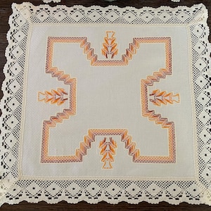 Può includere: Un centrino quadrato in tessuto bianco sporco con un motivo centrale a croce geometrica ricamato in tonalità arancione e marrone. Il pezzo è rifinito con un ampio e intricato bordo in pizzo all'uncinetto color crema con motivo a rombi.