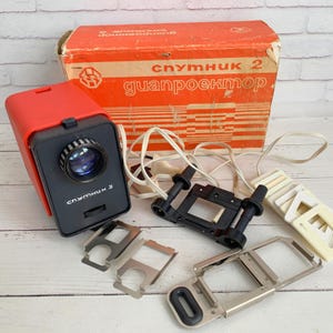 Puede incluir: Proyector de diapositivas rojo y negro vintage con el texto "Спутник 2". La imagen muestra el proyector, una caja con texto cirílico y varios accesorios, incluidos porta diapositivas y un cable de alimentación. Los objetos están sobre una superficie de madera blanca.