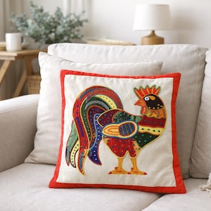 May include: A cushion with a vibrant rooster design. The rooster is depicted in red, orange, yellow, green, blue, and black. The cushion has a white background with a red border and is placed on a light-coloured sofa.
