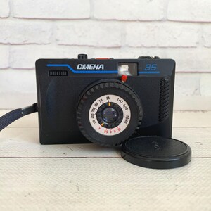 May include: Black Smena 35 camera with a blue strap and lens cap. The camera has the word "CMEHA" in blue and the number "35" on the front. The lens has a dial with numbers and markings.