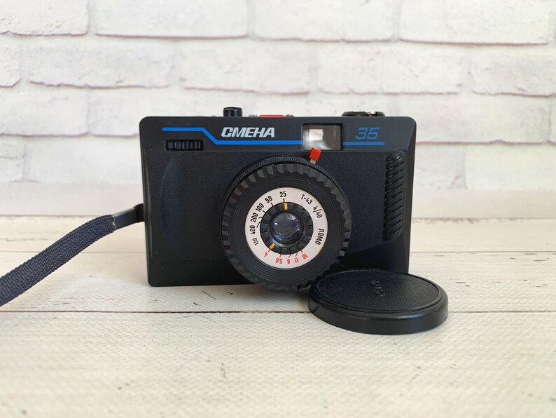 May include: Black Smena 35 camera with a blue strap and lens cap. The camera has the word "CMEHA" in blue and the number "35" on the front. The lens has a dial with numbers and markings.
