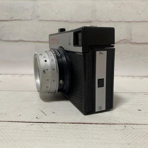 May include: A vintage silver and black camera with a lens and a textured body. The camera has a silver lens with numbers and markings. The camera is on a white surface.