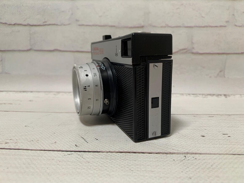 May include: A vintage silver and black camera with a lens and a textured body. The camera has a silver lens with numbers and markings. The camera is on a white surface.