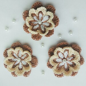 以下が含まれることがあります： 茶色とクリーム色の花びらと白いビーズの中心を持つ、手編みの花が3つあります。