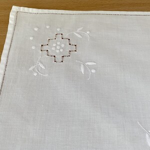 May include: A white square napkin featuring delicate white embroidery in the corner. The design includes a central geometric cross-like motif surrounded by small dots and curved leaf-like sprigs. The edges are finished with a hemstitched border.