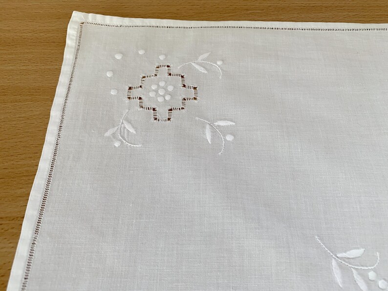 May include: A white square napkin featuring delicate white embroidery in the corner. The design includes a central geometric cross-like motif surrounded by small dots and curved leaf-like sprigs. The edges are finished with a hemstitched border.