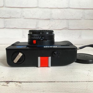 May include: Black OMEGA 35mm film camera with a black lens and a red shutter button. The camera has a black wrist strap and a black lens cap. The camera is on a white wooden surface.