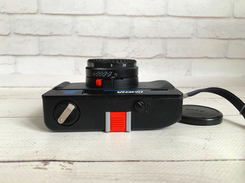 May include: Black OMEGA 35mm film camera with a black lens and a red shutter button. The camera has a black wrist strap and a black lens cap. The camera is on a white wooden surface.