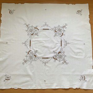 May include: A square white tablecloth featuring intricate brown and black floral embroidery in the center and at each corner. The fabric has a scalloped edge, and the central design consists of four large flowers connected by linear patterns.