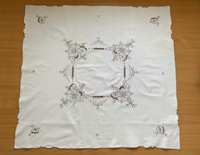 May include: A square white tablecloth featuring intricate brown and black floral embroidery in the center and at each corner. The fabric has a scalloped edge, and the central design consists of four large flowers connected by linear patterns.