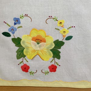 May include: Embroidered white linen table runner with a yellow scalloped edge. The centre features a large yellow flower with an orange centre, surrounded by green leaves, blue and red flowers.