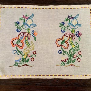 May include: A rectangular off-white linen placemat featuring two identical vertical floral embroidery patterns in blue, orange, green, and red. The border is finished with a decorative running stitch in alternating yellow and dark red thread.