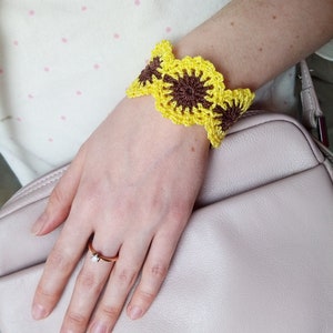 Puede incluir: Un brazalete de girasol tejido a crochet con hilo amarillo y marrón. El brazalete se lleva en la muñeca.