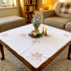 May include: A square, wooden coffee table with a white tablecloth featuring embroidered Christmas bells and bows. The table is decorated with a small Christmas tree, ornaments, and candles. The table is approximately 48 inches square.