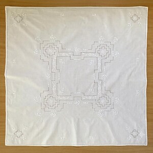 May include: A square white fabric napkin featuring intricate white embroidery. The center displays a geometric stepped pattern with small floral accents, while each of the four corners is decorated with a matching embroidered floral motif.