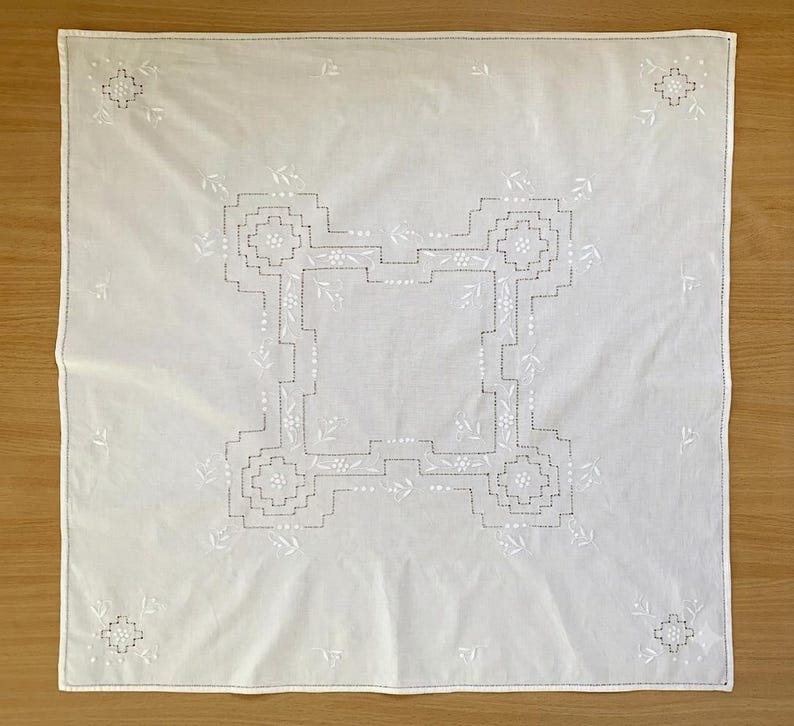 May include: A square white fabric napkin featuring intricate white embroidery. The center displays a geometric stepped pattern with small floral accents, while each of the four corners is decorated with a matching embroidered floral motif.
