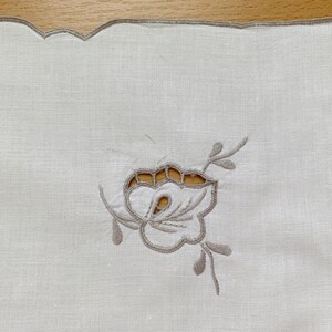 May include: A white rectangular linen napkin featuring a scalloped edge finished with light brown embroidery. The center displays a cutwork floral motif embroidered in light brown thread, showcasing delicate petals and leaves with open spaces.