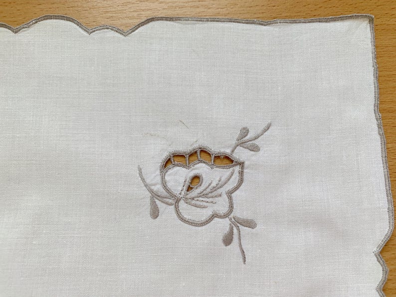 May include: A white rectangular linen napkin featuring a scalloped edge finished with light brown embroidery. The center displays a cutwork floral motif embroidered in light brown thread, showcasing delicate petals and leaves with open spaces.
