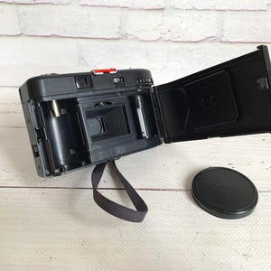 May include: Black vintage film camera with the back open, revealing the interior components. A black lens cap sits to the side. The camera has a red button and a black strap. The camera is on a white wooden surface.