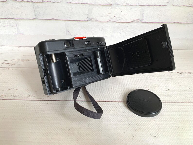 May include: Black vintage film camera with the back open, revealing the interior components. A black lens cap sits to the side. The camera has a red button and a black strap. The camera is on a white wooden surface.