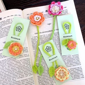 May include: Handmade crochet bookmarks with floral designs. Each bookmark features a colorful flower, green stem, and a tag that says "Bookmark". The bookmarks are placed on the pages of an open book.
