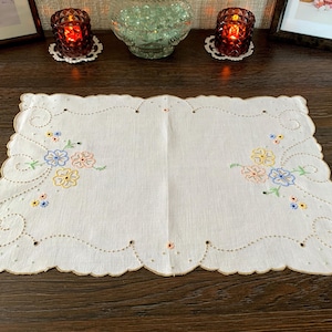 May include: A rectangular white placemat featuring scalloped edges with a tan border. The fabric is decorated with embroidered floral clusters in blue, yellow, and peach, accented by delicate green stems and intricate cutwork eyelet details.